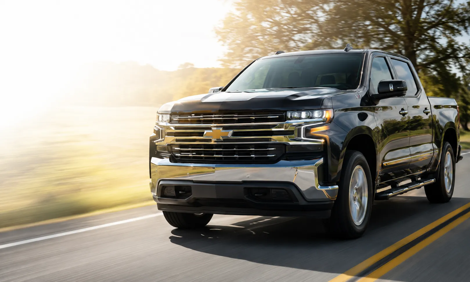 Black Chevrolet Silverado pickup driving on a sunlit road, front view with chrome grille and headlights visible